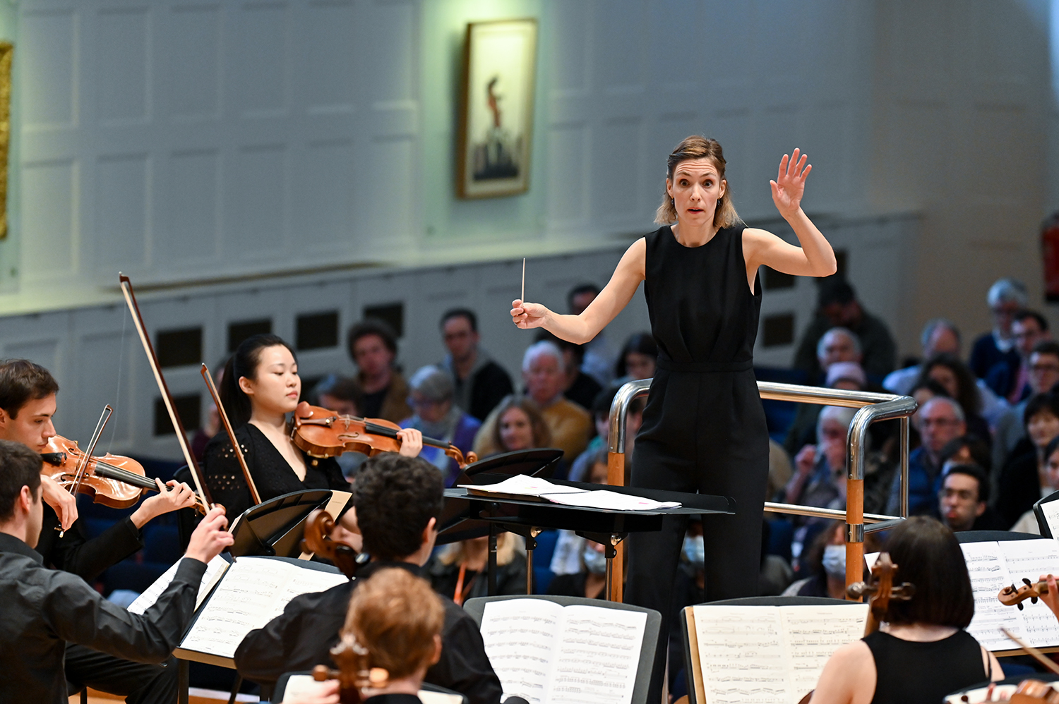 A female conductor dressed in black mid conducting in front of an orchestra test